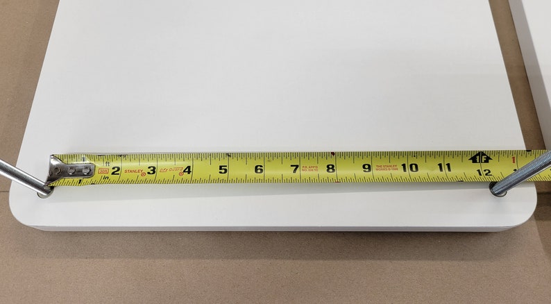 May include: A white shelf with a yellow tape measure showing a measurement of 11 1/2 inches. The shelf has two silver metal brackets attached to the underside.