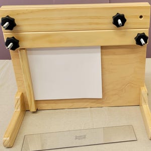 May include: A wooden printing press with a white paper sheet in the center. The press has two wooden arms with black knobs on each side. The press is sitting on a light brown surface.