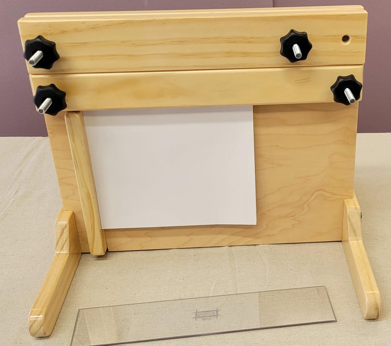 May include: A wooden printing press with a white paper sheet in the center. The press has two wooden arms with black knobs on each side. The press is sitting on a light brown surface.