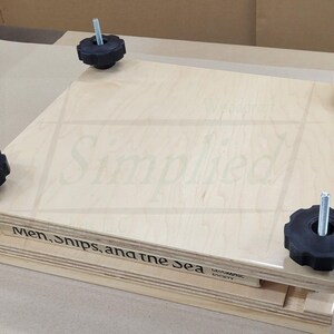 May include: A square wooden book press featuring two light-colored plywood boards held together by four metal threaded rods with black star-shaped knobs. The side of the bottom board displays the text "Men, ships, and the sea" and "210-1".