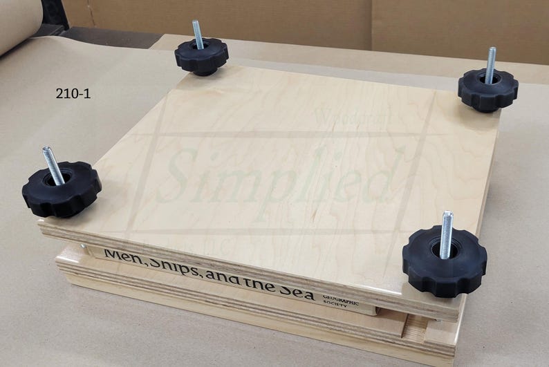 May include: A square wooden book press featuring two light-colored plywood boards held together by four metal threaded rods with black star-shaped knobs. The side of the bottom board displays the text "Men, ships, and the sea" and "210-1".