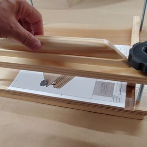 May include: A light-colored wooden book press featuring two horizontal boards connected by threaded metal rods and two black plastic adjustment knobs. A white instruction sheet with a technical diagram is placed between the wooden boards.