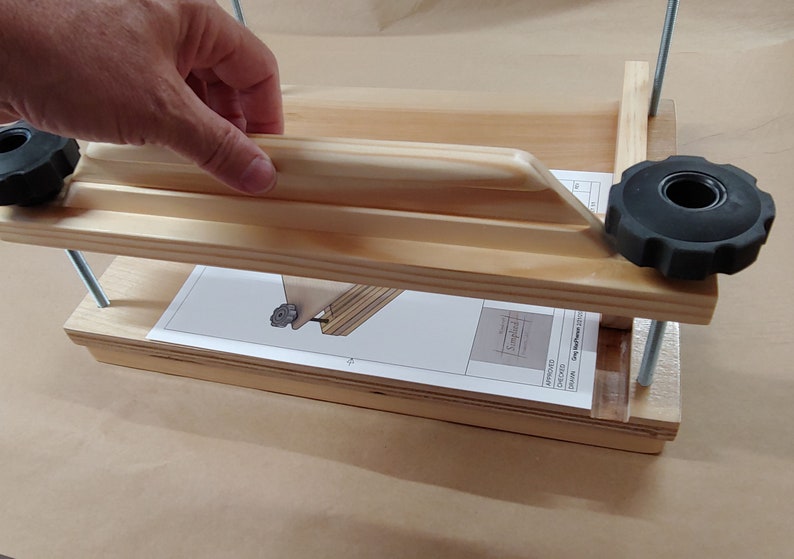 May include: A light-colored wooden book press featuring two horizontal boards connected by threaded metal rods and two black plastic adjustment knobs. A white instruction sheet with a technical diagram is placed between the wooden boards.