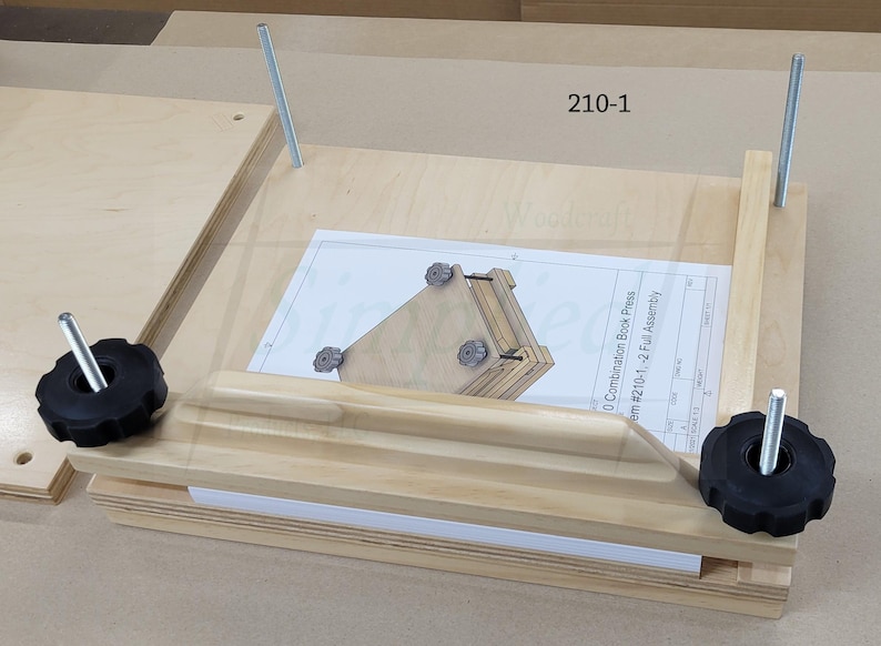 May include: A light-colored wooden book press featuring two vertical metal threaded rods and two black plastic star-shaped knobs. A white instruction sheet labeled 210-1 is placed on the base, showing a diagram of the assembly.