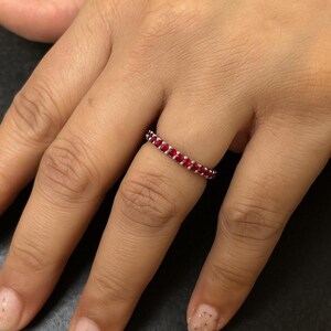 May include: A silver ring set with a continuous row of small, vibrant red gemstones. The ring is shown on a person's finger, with the gemstones catching the light. The ring is a classic design, suitable for everyday wear or special occasions.