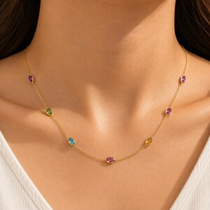 May include: A gold chain necklace featuring evenly spaced, bezel-set oval gemstones in various colors including purple, green, blue, pink, and yellow. The delicate metal chain connects each individual stone setting in a continuous, symmetrical design.