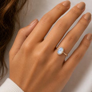 May include: A gold ring featuring a smooth, oval-shaped moonstone cabochon set in a simple metal bezel. The gemstone displays a soft, iridescent blue glow against the polished gold band, which rests on a finger, showcasing the ring's design.