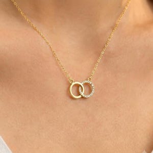 May include: A gold-toned necklace featuring two interlocking circular pendants. One circle is a plain polished gold band, while the other is a gold band encrusted with small, clear, sparkling stones. The pendant hangs from a delicate gold chain.