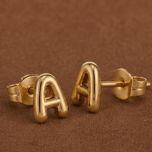 May include: A pair of gold-toned stud earrings shaped like the capital letter A. Each earring features a smooth, rounded, metallic finish with a post back closure. The design is simple and uniform, showcasing the letter A in a bold, sans-serif font.