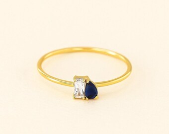 May include: A delicate gold ring featuring a rectangular clear stone and a teardrop-shaped dark blue stone. The ring has a thin, polished gold band, and the stones are set close together, creating a modern and elegant design. The ring is set against a soft, neutral background.
