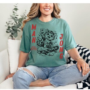 Può includere: T-shirt color verde acqua con un gatto dei cartoni animati su uno skateboard, che tiene una tessera del Mahjong. Le parole "MAH JONG" sono stampate verticalmente in rosso su entrambi i lati del gatto, con "READ BETWEEN THE TILES" sotto. La maglietta è di vestibilità ampia.