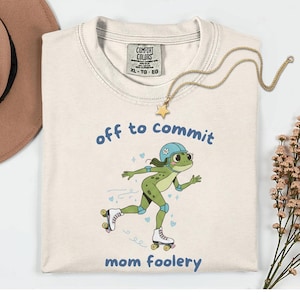 May include: Cream-coloured t-shirt with the text "off to commit mom foolery" and a cartoon frog on roller skates. The frog is green with a blue helmet and knee pads. A gold necklace with a star pendant is draped on the shirt.