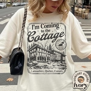 Puede incluir: Camiseta color marfil con el texto "I'm Coming to the Cottage" y un gr&aacute;fico de una caba&ntilde;a moderna. El gr&aacute;fico incluye el texto "Lanaudi&egrave;re, Qu&eacute;bec, Canada" y "Home to the Stupid Canadian Wolf Birds."