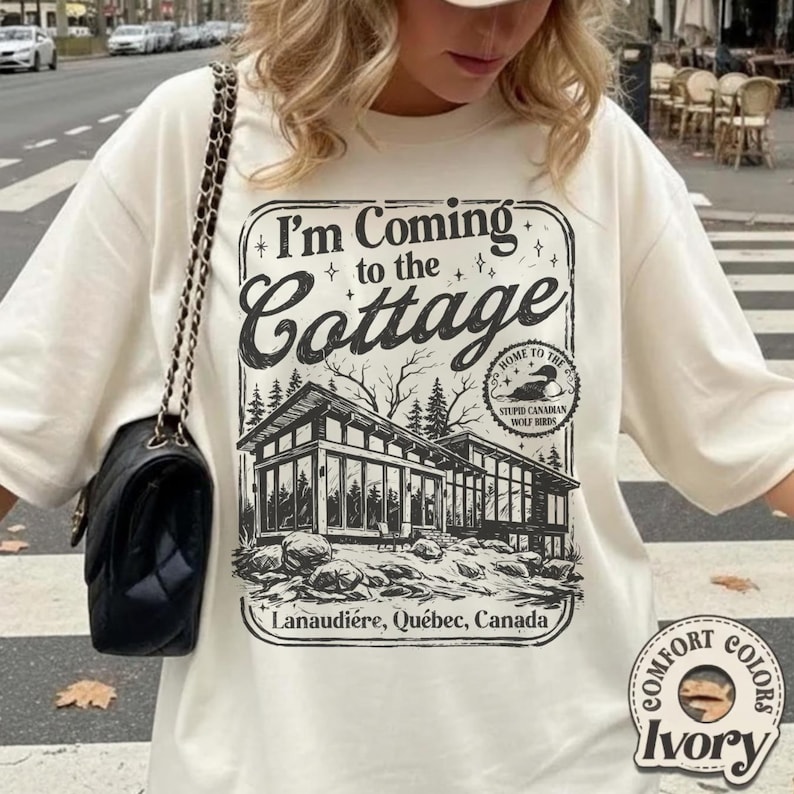 Puede incluir: Camiseta color marfil con el texto "I'm Coming to the Cottage" y un gr&aacute;fico de una caba&ntilde;a moderna. El gr&aacute;fico incluye el texto "Lanaudi&egrave;re, Qu&eacute;bec, Canada" y "Home to the Stupid Canadian Wolf Birds."