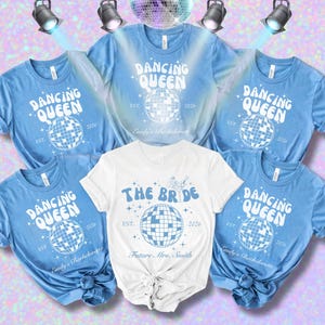 May include: Several light blue and white t-shirts featuring a disco ball design and the phrases "Dancing Queen" and "The Bride". The shirts are displayed against a pink and purple backdrop, with spotlights and a disco ball.