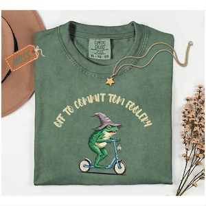 Può includere: T-shirt verde con la scritta "Off to Commit Tom Foolery" e una grafica di una rana su uno scooter blu, con un cappello da mago. Visibili anche una collana con pendente a stella dorata e un cappello marrone.