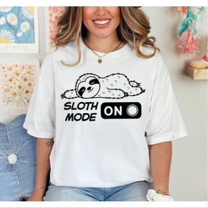 Può includere: T-shirt bianca con un'illustrazione in bianco e nero di un bradipo sdraiato. Il testo "SLOTH MODE ON" è stampato sotto la grafica del bradipo, con un design a interruttore "ON".