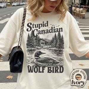 Può includere: T-shirt color avorio con una grafica raffigurante un'immersione in acqua, una baita e il testo "Stupid Canadian" e "WOLF BIRD". La maglietta ha anche un'etichetta "Comfort Colors".