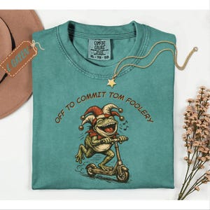 Può includere: T-shirt Comfort Colors verde acqua con una grafica di una rana su uno scooter e con un cappello da giullare. Il testo "OFF TO COMMIT TOM FOOLERY" è arcuato sopra la rana. Una collana con pendente a stella dorata e un cappello marrone sono anche nell'immagine.