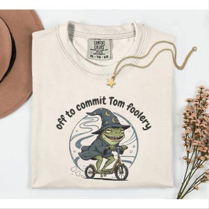 Könnte beinhalten: Cremefarbenes T-Shirt mit dem Aufdruck "off to commit Tom foolery" und einem Cartoon-Frosch mit Zaubererhut und Umhang, der Roller fährt. Eine goldene Halskette mit Sternanhänger und ein brauner Hut sind ebenfalls zu sehen.