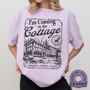 May include: A light purple t-shirt with a black graphic that reads "I'm Coming to the Cottage" above a detailed illustration of a modern cabin. The text "Lanaudière, Québec, Canada" is below the cabin image. The shirt is a Comfort Colors Orchid.