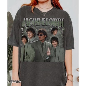 Può includere: T-shirt grigio scuro con la scritta "JACOB ELORDI" in un carattere verde stilizzato. La maglietta mostra più immagini di un uomo in giacca e cravatta e occhiali da sole. La parola "PEPPER" è stampata in basso a sinistra.