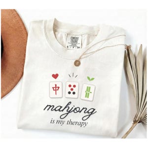 Può includere: T-shirt color crema con grafica di tre tessere di Mahjong, un cuore rosso e la scritta "mahjong is my therapy". Una collana d'oro con un ciondolo è appoggiata sulla maglietta. La maglietta è piegata e un cappello marrone è visibile sullo sfondo.