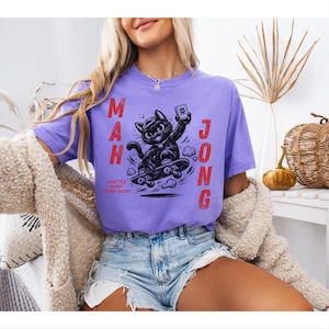 May include: A lilac-colored t-shirt featuring a black cat riding a skateboard and holding a Mahjong tile. The text "MAH JONG" is printed vertically on the left and right sides of the cat graphic. The phrase "ONE TILE AWAY FROM GLORY" is printed below the cat.