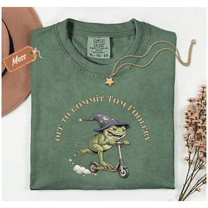 Könnte beinhalten: Moosgrünes T-Shirt mit dem Text "OFF TO COMMIT TOM FOOLERY", der eine Cartoon-Frosch auf einem Roller umgibt, der einen Zaubererhut trägt. Eine goldene Stern-Anhänger-Halskette und ein Etikett mit der Aufschrift "Moss" sind ebenfalls sichtbar.