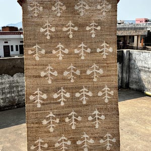 May include: A rectangular, woven jute rug with a repeating pattern of white, stylized tree-like shapes. The rug is held up, showcasing its natural brown color and textured surface. The design is symmetrical and suitable for home decor.