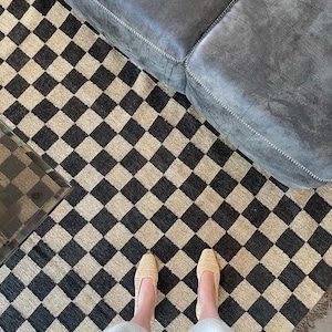 Peut inclure: Vue aérienne d'un tapis à carreaux noirs et beiges avec un canapé gris en arrière-plan. Des chaussures beiges tissées sont visibles sur le tapis. Le tapis a une bordure frangée.