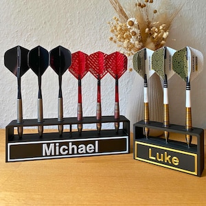 Könnte beinhalten: Zwei schwarze Dartständer aus Kunststoff mit Dartpfeilen. Der größere Ständer hält sechs Pfeile mit schwarzen und roten Flights und trägt den Namen Michael. Der kleinere Ständer hält drei Pfeile mit gemusterten weißen Flights und den Namen Luke.