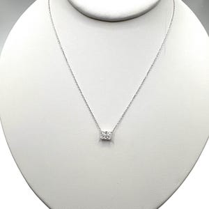 Puede incluir: Un delicado collar de plata con un colgante rectangular. El colgante está adornado con pequeñas piedras brillantes. El collar se exhibe sobre un busto blanco, resaltando su elegante diseño. La cadena es fina.