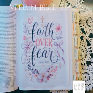 May include: A translucent vellum insert for a Bible featuring the hand-lettered text "faith OVER fear" and "ISAIAH 41:10" in dark purple and pink. The design includes a cross, hearts, floral illustrations, and ribbons, with adhesive tabs attached to the side.