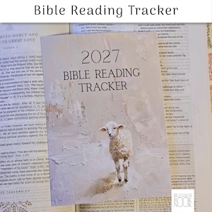 May include: A 2027 Bible Reading Tracker booklet featuring a textured, off-white painted background with a small lamb illustration. The text 2027 BIBLE READING TRACKER is printed in black on the front cover, which rests atop an open book page.