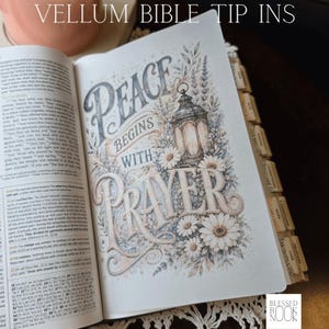 May include: A vellum bible tip-in page featuring the text Peace Begins With Prayer in decorative lettering, accompanied by an illustration of a lantern and white daisies. The page is inserted into a bible with visible index tabs on the side.