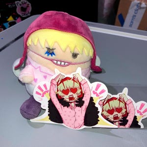 May include: A plush doll with a pink hat and pajamas, and a sticker featuring an anime character with heart-shaped eyes. The doll has yellow hair and a stitched-on face. The sticker has a clear border.