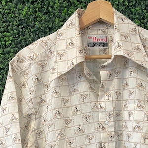 May include: Cream-colored button-down shirt with a repeating brown bicycle pattern. The shirt has short sleeves and a classic collar. The brand name "Breed" is visible on the label.