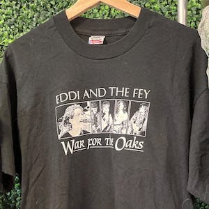 May include: Black t-shirt with the text "EDDI AND THE FEY" above a graphic of five figures and the words "War for the Oaks" in a stylized font. The shirt is hanging on a wooden hanger against a green background.