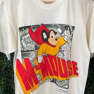 Puede incluir: Camiseta de color crema con un panel de cómic con el personaje Mighty Mouse. El panel incluye el texto "Mighty Mouse" en letras grandes, rojas y amarillas. El personaje está representado con una capa roja y una camisa amarilla.
