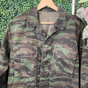 May include: A vintage tiger stripe camouflage jacket with four front pockets and a button-up closure. The jacket features a green, brown, and black pattern. The jacket is hanging on a wooden hanger.