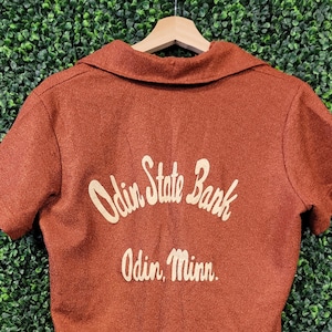 May include: A short-sleeved, rust-colored shirt with a collared neckline. The back of the shirt features the text "Odin State Bank" above "Odin, Minn." in a cream-colored, stylized font. The shirt is hanging on a wooden hanger against a green background.