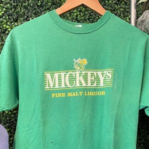 May include: A green t-shirt featuring the text "MICKEY'S FINE MALT LIQUOR" in yellow. The shirt has a crew neck and short sleeves. The graphic includes a cartoon bee. The t-shirt is displayed on a wooden hanger.
