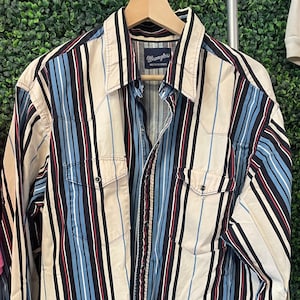 May include: A long-sleeve button-down shirt by Wrangler featuring a vertical striped pattern in cream, black, light blue, and red. The shirt includes a pointed collar, two chest pockets with snap closures, and a front button placket.