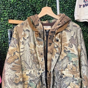 May include: A camouflage jacket with a hood and zipper closure. The jacket features a brown and green leaf pattern. It has two front pockets and brown ribbed cuffs and hem. The jacket is hanging on a wooden hanger against a green background.