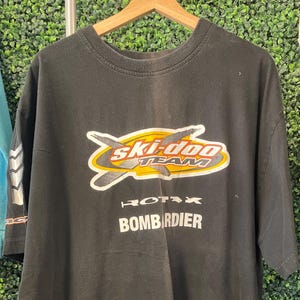 May include: Black t-shirt with a graphic that reads "Ski-Doo Team" in yellow, orange, and white. Below the logo are the words "Rotax" and "Bombardier" in white. The shirt is hanging on a wooden hanger.
