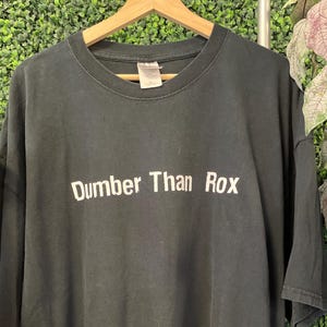 Puede incluir: Camiseta negra con el texto "Dumber Than Rox" en letras blancas. La camiseta está colgada de una percha de madera sobre un fondo verde y blanco. La camiseta es de estilo cuello redondo estándar.