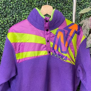 May include: A purple fleece pullover with a high collar and snap closure. The upper chest features a design with neon green and orange accents, along with the word "Nordica". The trim is neon green.