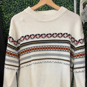 May include: A cream-colored long-sleeve knit sweater featuring a ribbed crew neckline, cuffs, and hem. The chest and sleeves are decorated with horizontal bands of geometric patterns in shades of dark red, black, grey, and mustard yellow.