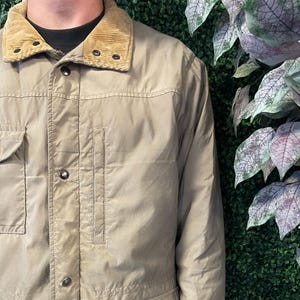 May include: A tan jacket with a corduroy collar. The jacket has four front pockets, a button-up closure, and a vertical pocket on the chest. The jacket is a neutral color and appears to be made of a durable material.